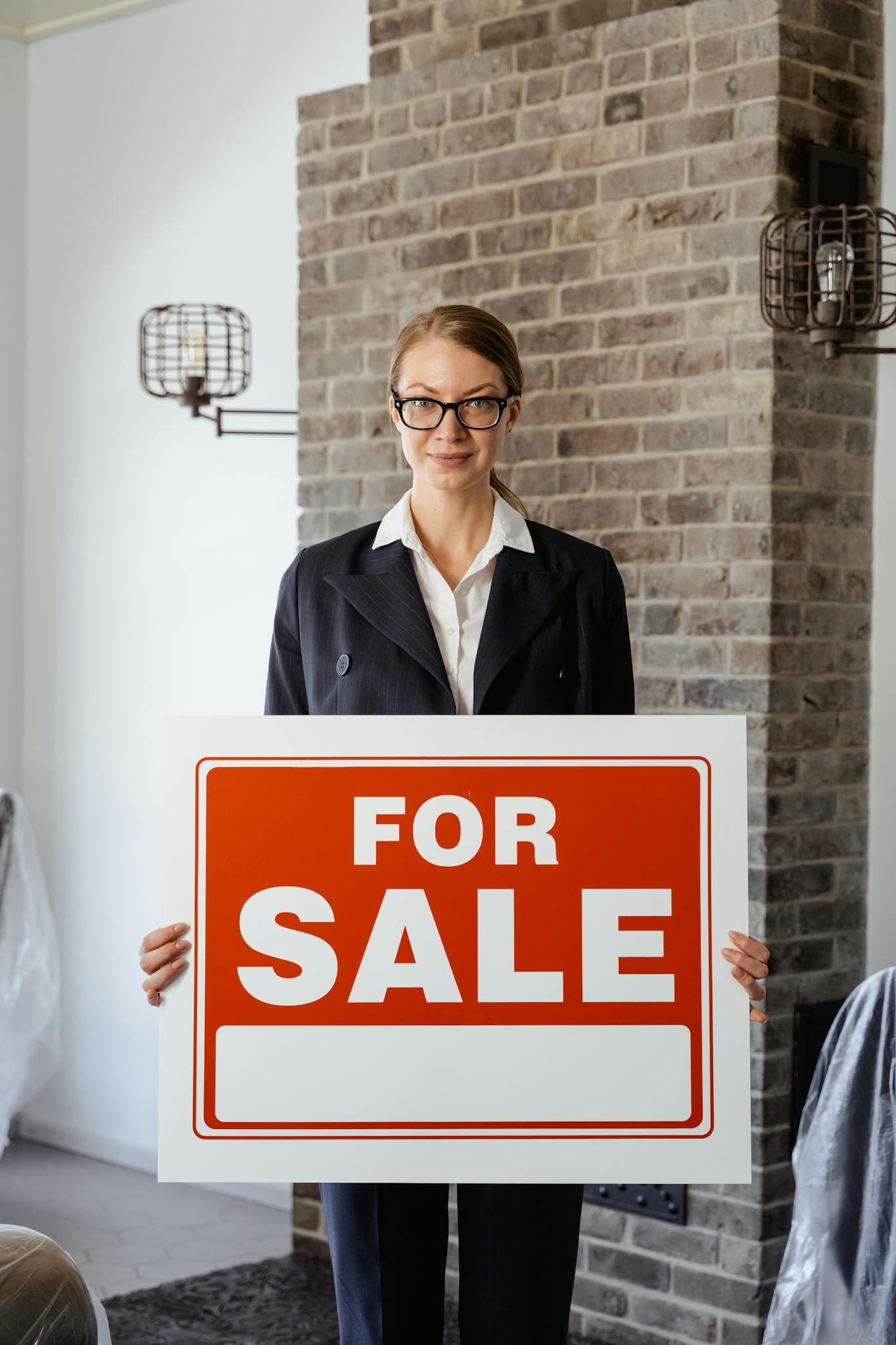 Female real estate agent holding a 'For Sale' sign inside, ready to assist clients.